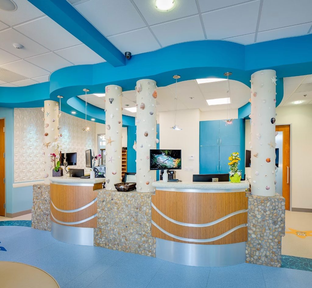 Curved wood and river-stone check-in desks at CarlsbaDDS Pediatric Smiles, framed by seashell-studded columns